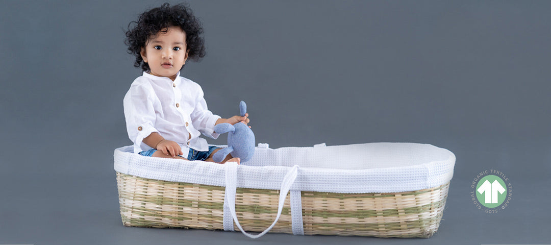 A baby in white organic cotton sitting in a woven moses basket, beside the GOTS certified organic textile mark